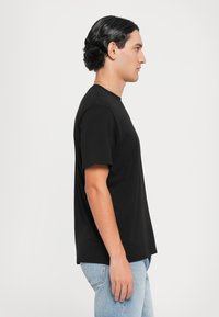 Svart kortermet T-skjorte laget av bomull, med rund hals og rett passform, brukt sammen med lyseblå jeans, vist fra siden.