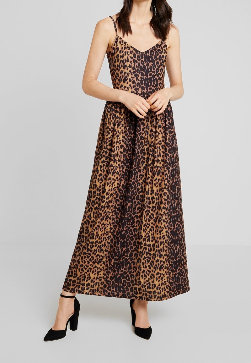 Robe longue à imprimé léopard avec fines bretelles, décolleté en V, tissu fluide et palette de couleurs marron et noir. Portée avec des chaussures à talons noires.