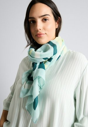 Jeune femme aux cheveux foncés portant un chemisier rayé clair et une grande écharpe à carreaux pastel bleu et vert enroulée autour du cou.