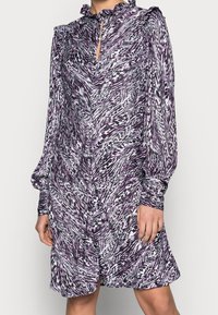 Fioletowa i biała sukienka w wzory z długimi rękawami, falbaniastym kołnierzem i detalem w kształcie dziurki. Wykonana z lekkiego materiału o swobodnym kroju.