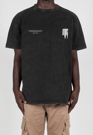 Mann trägt ein ausgewaschenes schwarzes übergroßes T-Shirt mit kleinen weißen Text- und Grafiklogos auf der Brust, kombiniert mit beigen Cargo-Hosen.