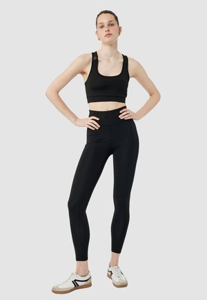 Vrouw staat met handen op de heupen, gekleed in een zwarte sport-bh, hoge zwarte legging en witte sneakers tegen een effen achtergrond.