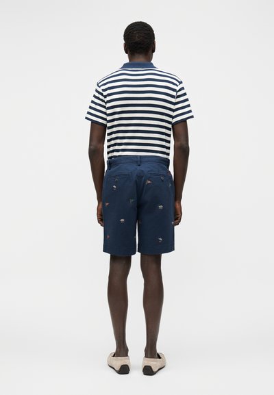 Άνδρας που φοράει σορτς σε ναυτικό μπλε χρώμα με κεντημένες ναυτικές σημαίες και λευκό μπλε ριγέ κοντομάνικο πουκάμισο, στέκεται με την πλάτη προς το μέρος μας.