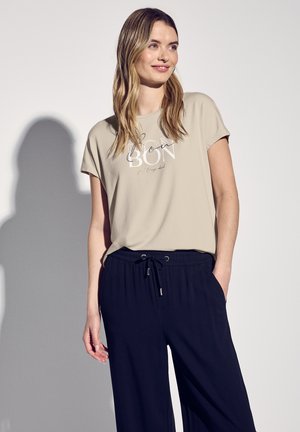 T-shirt beige à manches courtes, col rond, avec un texte imprimé blanc. Associé à un pantalon ample bleu marine avec une taille à cordon.