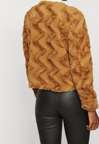 Brun faux fur-jacka med zigzag-textur, långa ärmar och kort design. Matchas med svarta, slim-fit byxor.