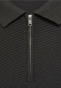 Haut polo noir en maille avec une fermeture éclair et un motif texturé. Le col est uni, contrastant avec la maille ajourée du corps.