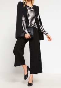 Czarna peleryna wierzchnia na paski z długim rękawem, zestawiona z czarnymi spodniami o szerokich nogawkach. Całość dopełnia czarna kopertówka i czarne baleriny.