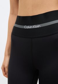 Primo piano di una persona che indossa leggings neri Calvin Klein con una fascia larga in vita caratterizzata da una striscia grigia e dal testo del logo.