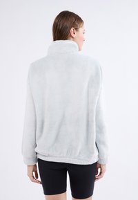 Veste en polaire gris clair doux avec un col haut, un ourlet élastique et des manches longues, présentant une texture pelucheuse et une coupe décontractée.