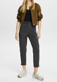 Femme portant une veste bomber olive, un top noir, un pantalon taille haute à cordon noir, et des baskets blanches se tenant devant un fond blanc.