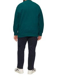 Un sweat-shirt vert avec un col rond et des poignets côtelés, associé à un pantalon sombre et des chaussures blanches avec un accent bleu.