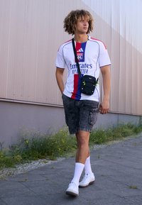 Maillot de football blanc avec des rayures rouges et bleues, shorts cargo gris, sac bandoulière noir et baskets blanches avec semelles grises, marchant sur le pavement.