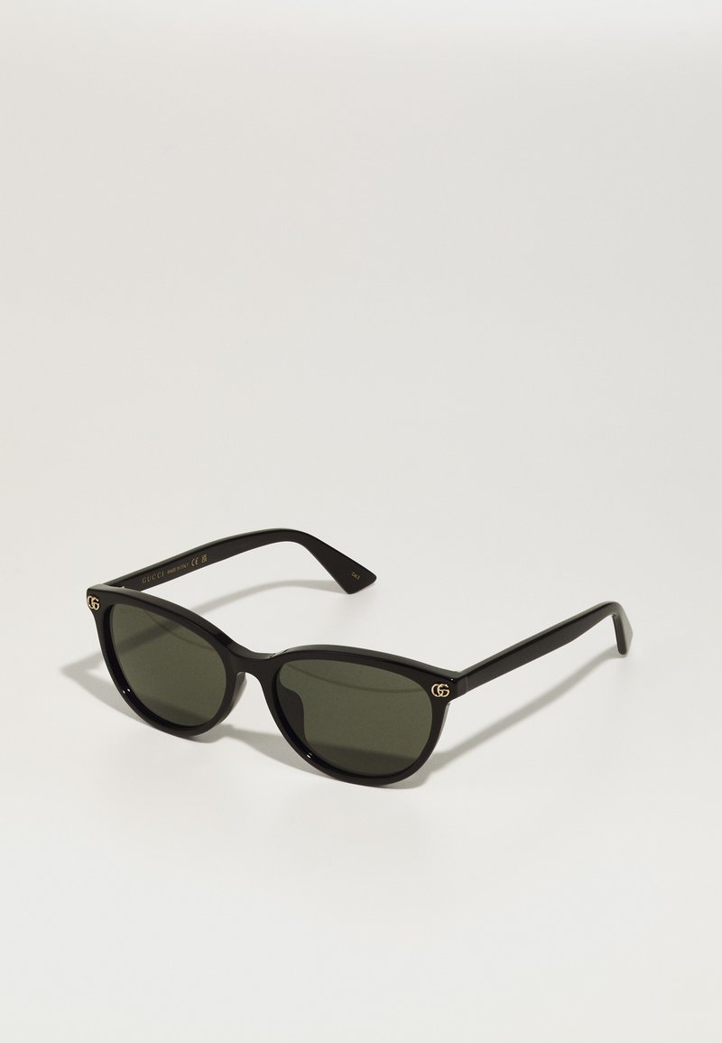Ochelari de soare cu un cadru din plastic negru, lentile verde închis și accente aurii cu logo-ul GG în față. Design subțire, unghiular, incluzând brațe drepte.