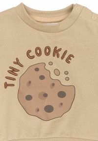 Béžová mikina s grafikou sušenky s hnědými skvrnami a vyšitým textem "TINY COOKIE". Kolo výstřih s druky na rameni.