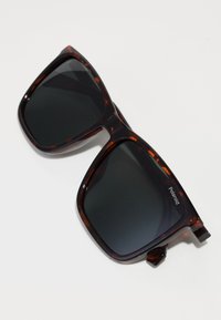 Ochelari de soare cu montură din scoață de broască țestoasă, cu lentile închise, formă pătrată, cadru din plastic transparent și marcaj Polaroid proeminent pe lateral.