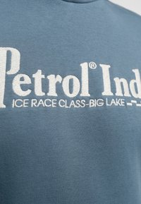 Světlě modrá mikina s bílým vyšitým nápisem "Petrol Ind" a "ICE RACE CLASS-BIG LAKE", s hladkou a měkkou texturou.