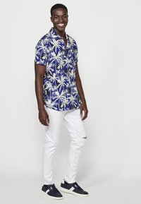 Camicia azzurra a maniche corte con motivo di palme bianche, abbinata a jeans bianchi strappati e sneakers blu marina con accenti bianchi.