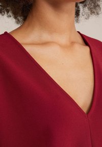 Burgundské šaty s výstřihem do V, které mají hladkou matnou texturu a čisté švy. Výstřih je ostrý, což zdůrazňuje elegantní design.