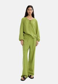 Zielony dwuczęściowy zestaw składający się z luźnej bluzki z wiązaniem oraz spodni z elastycznym paskiem. Gładka tkanina o miękkiej fakturze. Czarne sandały.