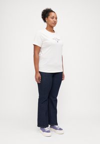 T-shirt en coton blanc à manches courtes, avec un texte logo rouge. Associé à un jean trapèze bleu foncé et des baskets violettes.