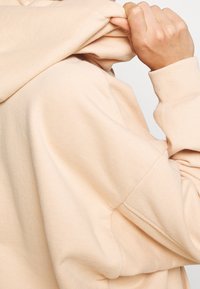 Felpa beige chiaro realizzata in tessuto morbido. Presenta una tasca a marsupio e polsini a coste. Primo piano di una mano che aggiusta il cappuccio.