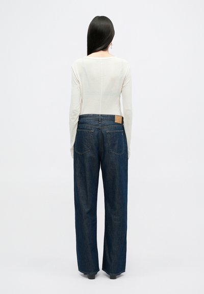 Lange mouwen off-white top gecombineerd met donkerblauwe wijde jeans met achterzakken en een gestructureerd tan leren label aan de taille.