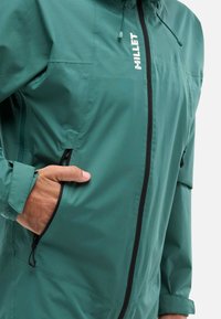 Veste en millet teal avec une texture légère et lisse. Dotée d'une fermeture éclair à l'avant, de poches latérales et d'une capuche avec cordon de serrage ajustable.