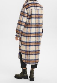 Person in einem langen, flauschigen karierten Mantel in Beige, Blau und Braun, kombiniert mit schwarzen, verkürzten Hosen und olivgrünen, klobigen Stiefeln.
