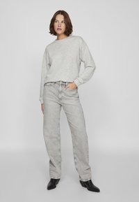 Mujer con cabello castaño corto, vistiendo un sudadera gris claro, jeans gris claro de tiro alto y botines negros, de pie con una mano en el bolsillo.