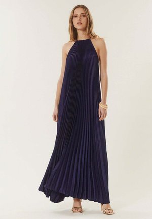 Femme portant une robe longue plissée sans manches bleu marine et des sandales dorées, debout devant un arrière-plan clair uni.