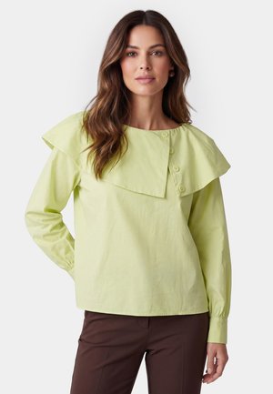 Woman with long brown hair wears light green blouse with large asymmetrical collar and buttons, paired with dark brown pants.