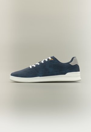 Marineblauer Sneaker in niedriger Ausführung mit weißen Schnürsenkeln, perforierten Seitenpaneelen, beigem Fersenlasche und weißer Gummisohle vor neutralem Hintergrund.