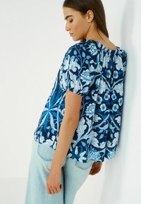 Blusa floreale blu con maniche a sbuffo, scollatura arricciata e un motivo testurizzato in diverse tonalità di blu su uno sfondo blu scuro. Indossata con jeans chiari.