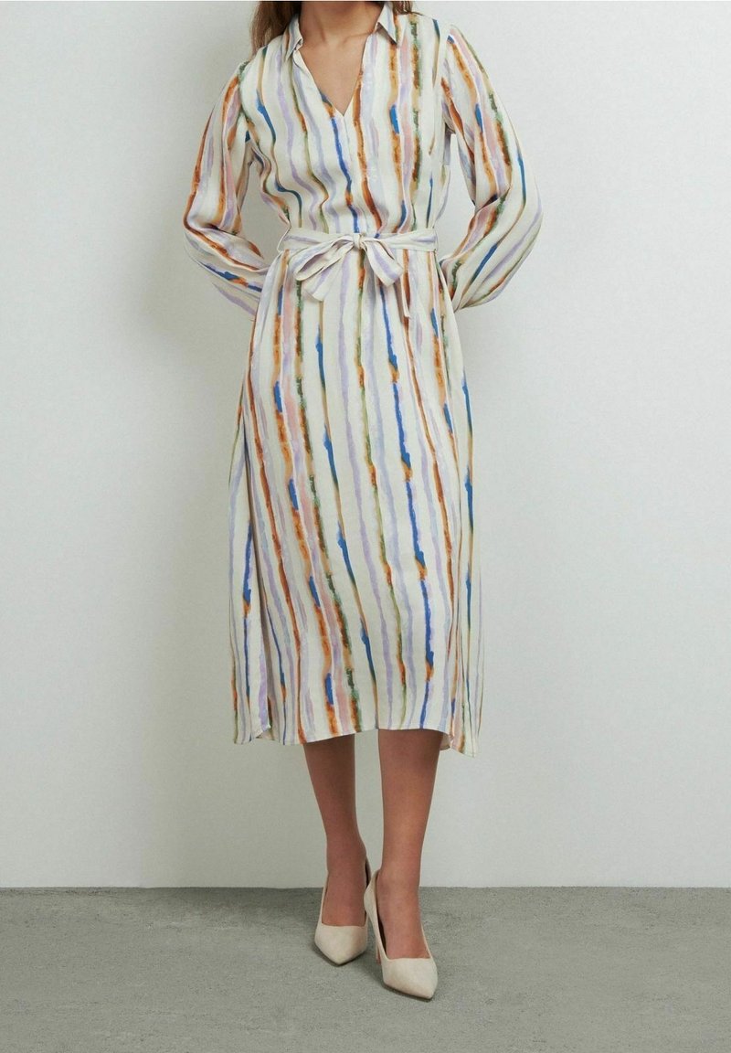 Robe à manches longues avec col et ceinture à la taille. Tissu blanc orné de fines rayures verticales multicolores en orange, bleu et vert. Longueur mi-mollet.