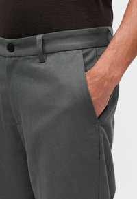 Szare spodnie z bawełny w połączeniu z innymi materiałami o gładkiej fakturze, prostym kroju i bocznymi kieszeniami. Posiadają zapięcie na guzik oraz szlufki na pasek w talii.