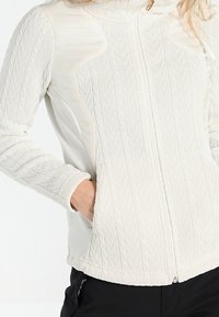 Giacca a zip di un beige chiaro con texture a trecce sulle maniche e sul davanti, tessuto liscio sui lati e colletto alto.