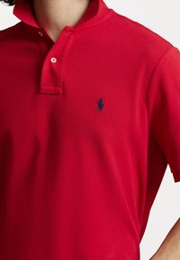 Červená polo tričko z texturované tkaniny, s límcem na knoflíky a námořnickým logem na levém horním rohu. Krátké rukávy, přiléhavý design.