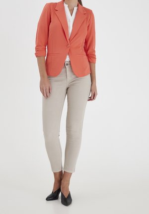 Frau mit korallenfarbener Blazer über einer weißen Bluse, beigen schmalen Hosen und schwarzen spitzen Absätzen, vor einem einfarbigen weißen Hintergrund stehend.