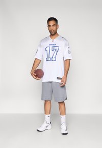 Balta futbola kreklā ar zilām detaļām un numuru 17, kopā ar pelēkām biksītēm. Modelis tur brūnu futbolbola bumbu, valkājot baltus sporta apavus.