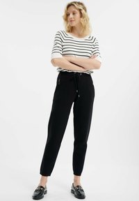 Schwarze Jogginghose mit Reißverschlusstaschen, kombiniert mit einem weiß gestreiften Shirt. Das Material wirkt weich und bequem. Die Beine sind schmal zulaufend.