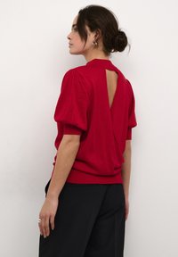 Pulover roșu cu mâneci bufante, design cu spate deschis și tiv cu nervuri. Textură tricotată cu un ușor strălucire. Asortat cu pantaloni negri.
