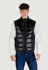 Chaleco negro acolchado con un acabado brillante, diseño de cuadros y cremallera frontal. Llevado sobre una camiseta de manga larga blanca, combinado con jeans azules.