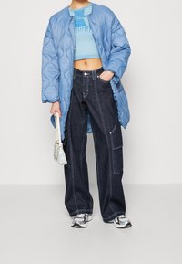 Puffet lyseblå jakke, cropped blå sweater, mørke løse jeans med hvide syninger. Tilbehør inkluderer en lille, struktureret taske og grå sneakers.