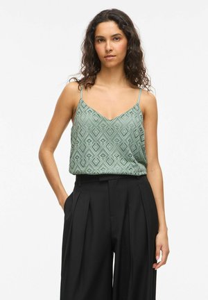 Femme aux cheveux bouclés et foncés portant un haut sans manches vert à motifs et un pantalon plissé noir taille haute, debout avec une main dans la poche.