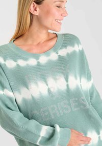 Mintgroene ribbeltrui met een witte tie-dye patroon en versierde tekst "ROCK & CERISES." Wijde halslijn en een loose fit.