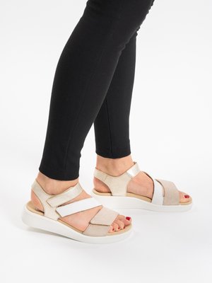 Voeten met beige-witte verstelbare sandalen met open tenen en dikke witte zolen, gecombineerd met zwarte nauwsluitende broek.