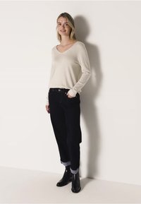 Světlý béžový svetřík s dlouhými rukávy a výstřihem do V, spárovaný s tmavými džínami, s ohrnutými manžetami a černými kotníkovými botami. Jednoduchý design, měkká textilie.