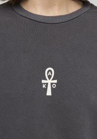 Sudadera gris oscuro con cuello redondo acanalado y un símbolo ankh blanco con "A" y "K" en el interior, situado en la zona del pecho.