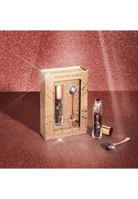 Set di occhi in ametista inserito in una scatola dorata; include un applicatore a rullo trasparente e un applicatore curvo con un'estremità viola, il tutto contro uno sfondo luccicante.