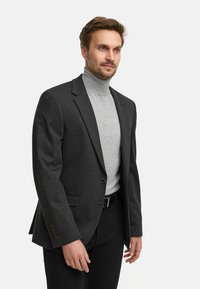 Mørk grå skreddersydd blazer med skjærte krager, to knapper og sidelommer. Brukt over en lys grå turtleneck og svarte bukser.